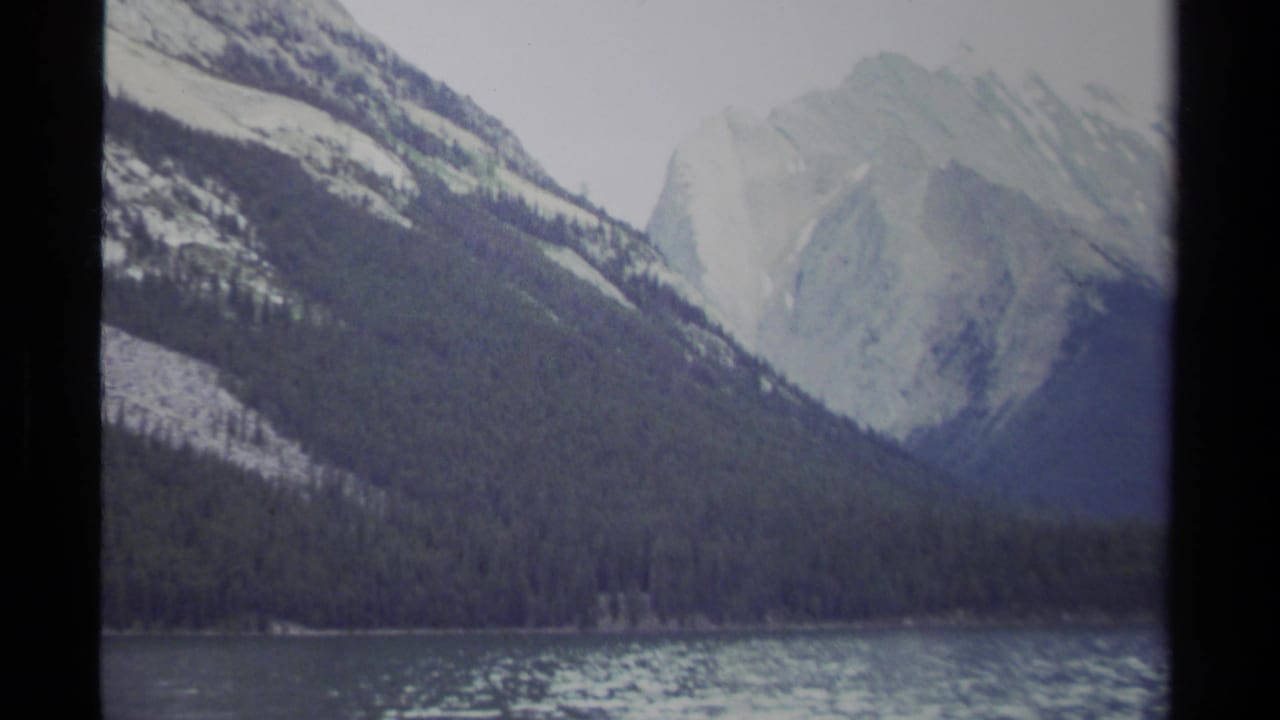1979 British Columbia 8mm frame showing a mountain lake, evergreen slopes, and pale snow-dusted peaks under a cloudy sky.