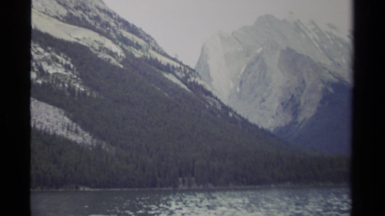 Similar mountain lake view with evergreen forest, rocky peaks, and visible film gate border.