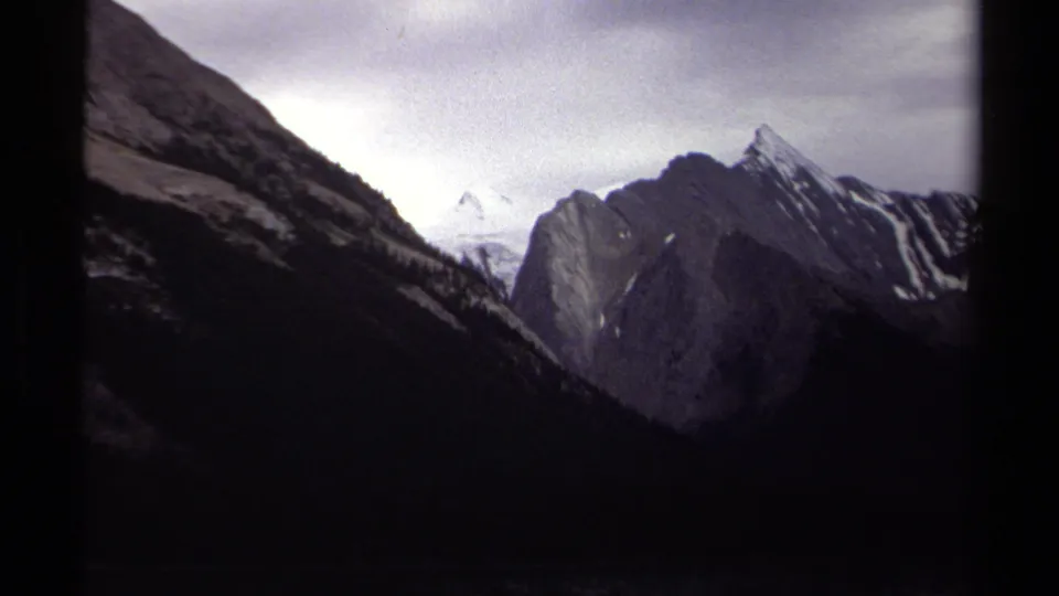 1979 British Columbia 8mm frame showing a darker mountain valley view with rocky slopes, snow patches, and distant pale peaks.