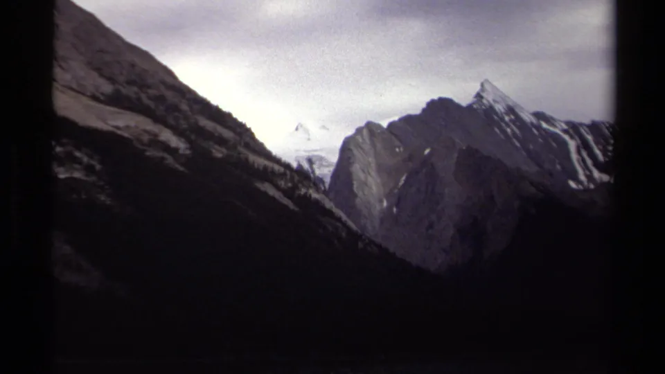 1979 British Columbia home-movie frame of a rugged mountain valley, with a pointed peak, shadowed slopes, and cloudy sky.