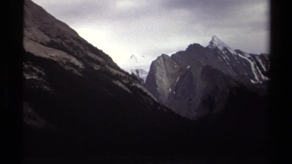 1979 British Columbia 8mm frame showing a dark alpine valley, steep ridges, and distant snow-covered mountains under cloudy sky.