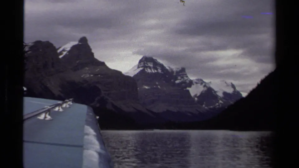 1979 British Columbia travel view across a lake toward rugged, partly snow-covered mountains, filmed from a vehicle or boat edge.