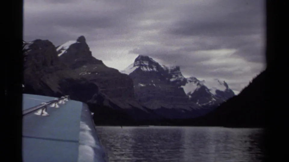 Vintage 8mm view across a British Columbia lake toward dark alpine ridges and snow-streaked peaks from a traveler's viewpoint.