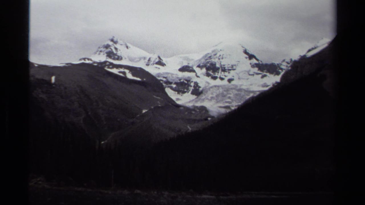 1979 British Columbia 8mm frame showing snow-covered alpine peaks beyond dark valley slopes and evergreen forest.