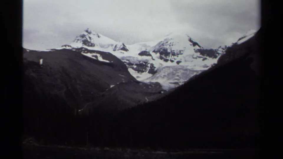 1979 British Columbia 8mm frame showing snow-covered alpine peaks beyond dark valley slopes and evergreen forest.