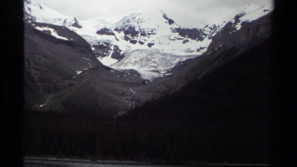 1979 British Columbia 8mm frame with snow-bright peaks, a central glacier or snowfield, and a dark evergreen foreground.
