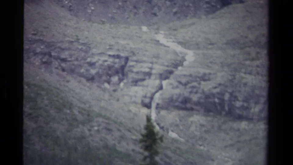 1979 British Columbia 8mm view of a rocky mountainside where a thin stream descends behind a small dark conifer.