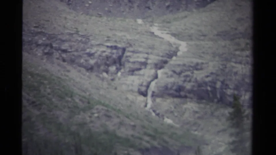 1979 British Columbia 8mm frame of a pale stream tracing down a grey rocky slope with soft, blurred vegetation in front.
