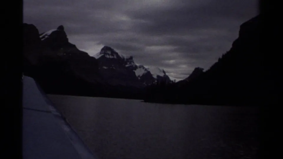 1979 British Columbia 8mm lake landscape with dark mountain silhouettes, snow-touched peaks, and a heavy overcast sky.