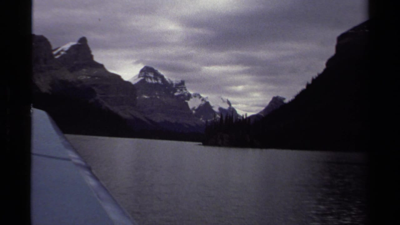 Boat-level view across a lake toward snow-streaked mountains and dark forest under cloudy sky.