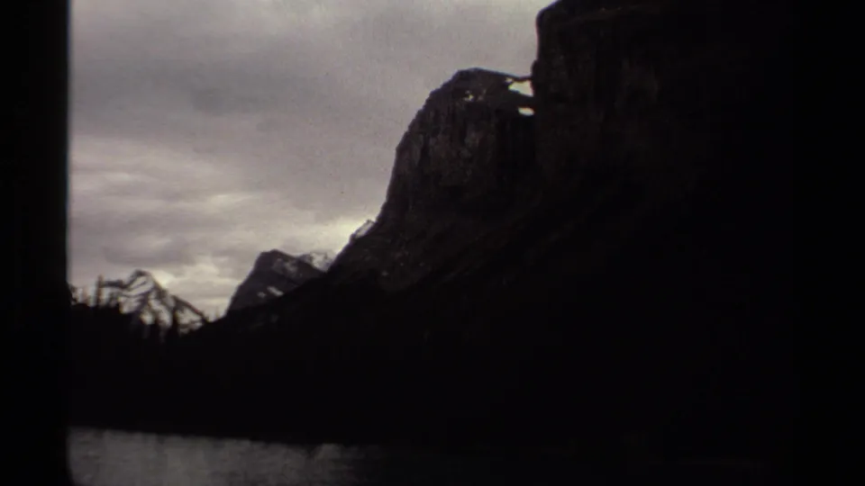 1979 British Columbia 8mm frame with a dark rock face, lake edge, distant peaks, and heavy cloud cover from a boat perspective.