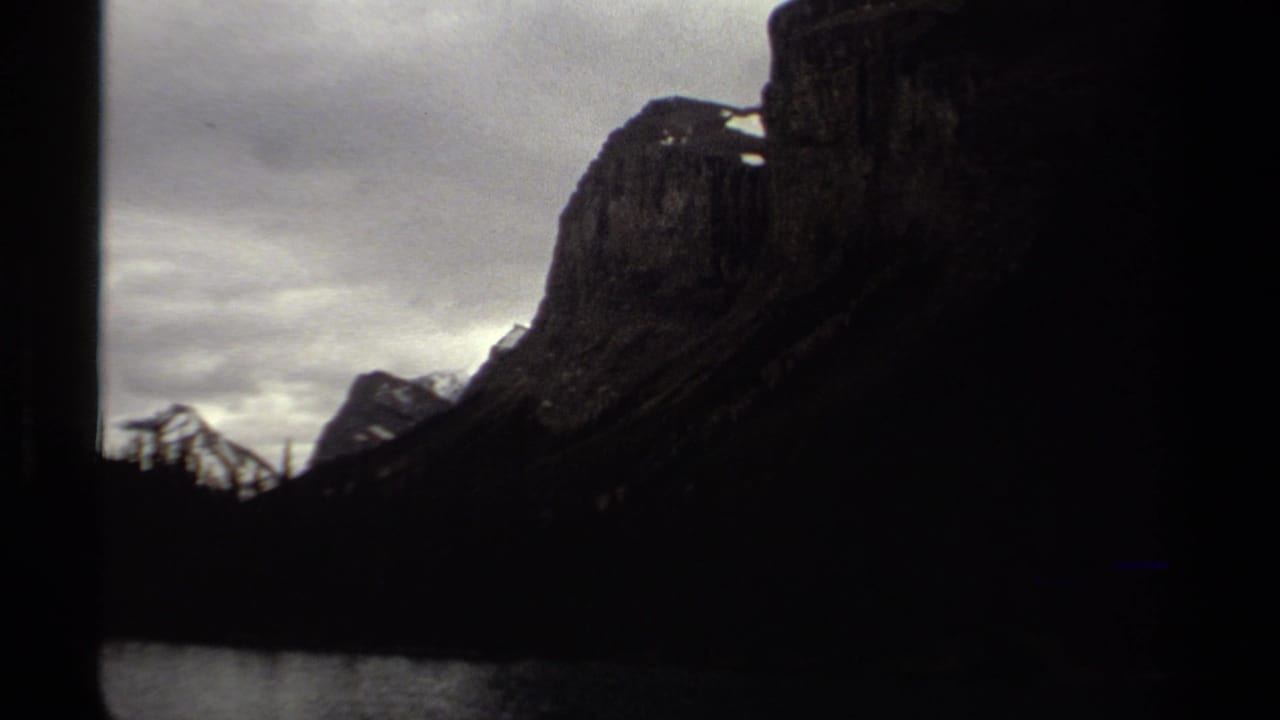 British Columbia 1979 8mm frame showing a dark mountain lakeshore under gray clouds, with soft grain and black frame edges.