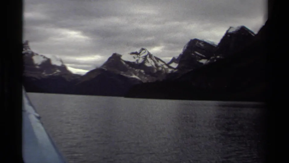 British Columbia 1979 8mm travel frame with the boat edge at left, rippled lake water, and dark snowy mountains in the distance.