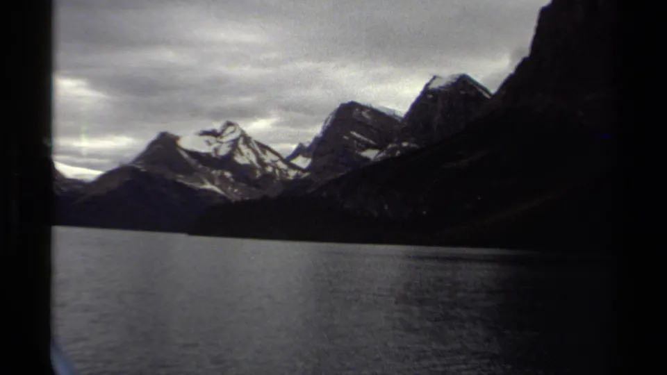 1979 British Columbia 8mm frame of a quiet mountain lake bordered by dark slopes and snow-streaked peaks beneath overcast skies.