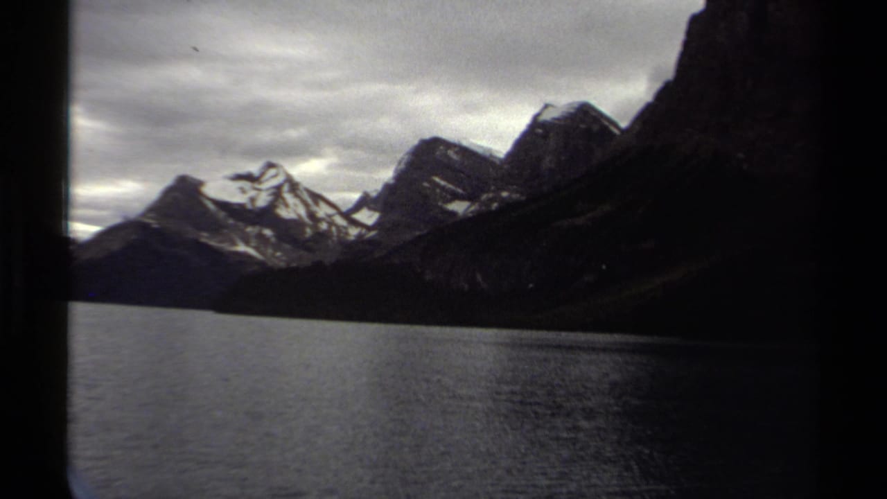Calm lake below dark mountain slopes and snow-dusted peaks under a cloudy sky.
