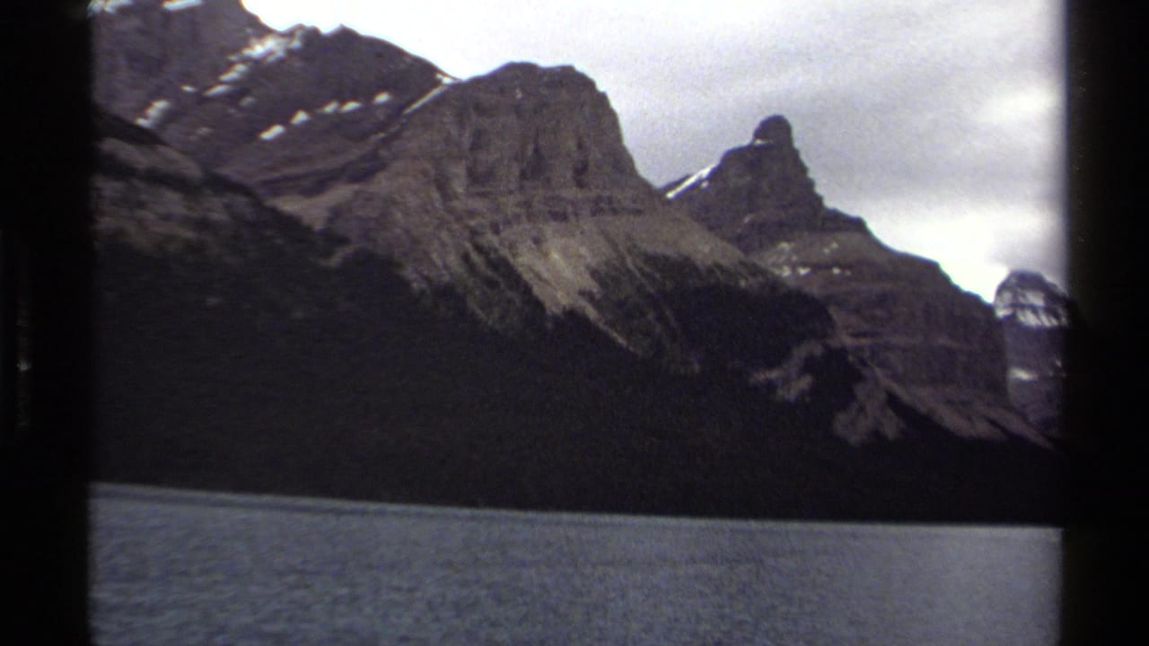 Rocky mountains with snow patches rise above a lake under an overcast sky.