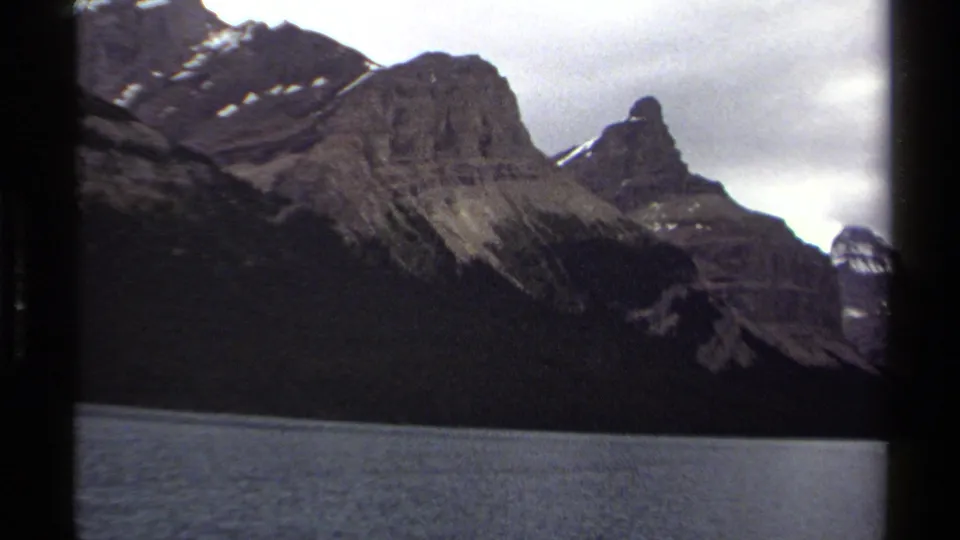1979 British Columbia 8mm frame showing rugged mountain peaks, dark forested slopes, and calm lake water beneath a cloudy sky.