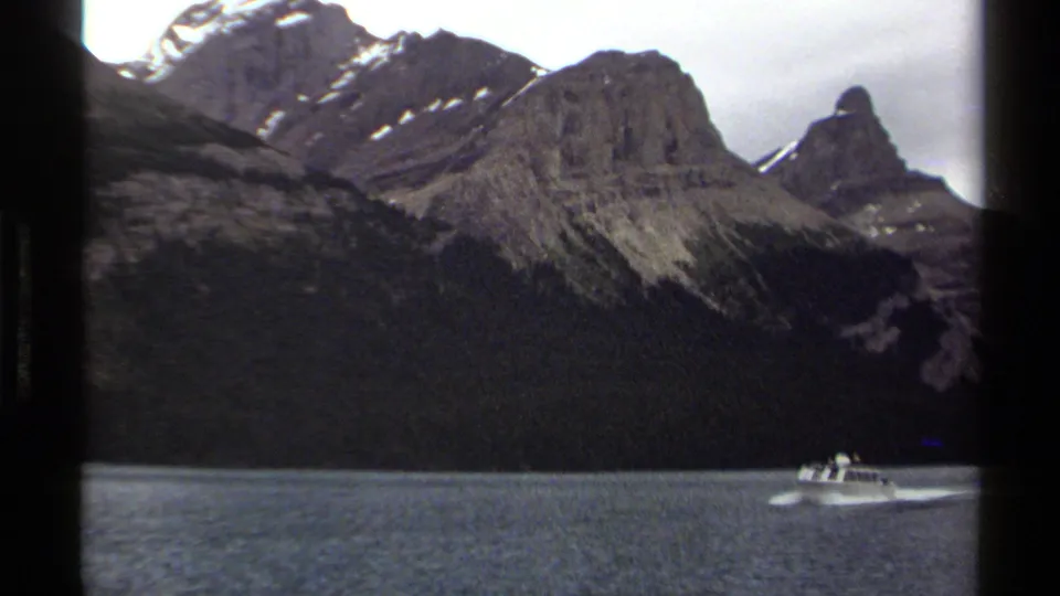 1979 British Columbia 8mm frame with a white motorboat and distant occupants moving across a lake below rugged mountains.
