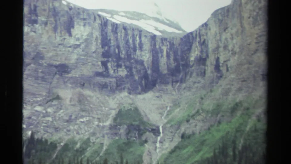 1979 British Columbia 8mm frame showing a steep green valley, pale rock faces, snow remnants, and a narrow waterfall.