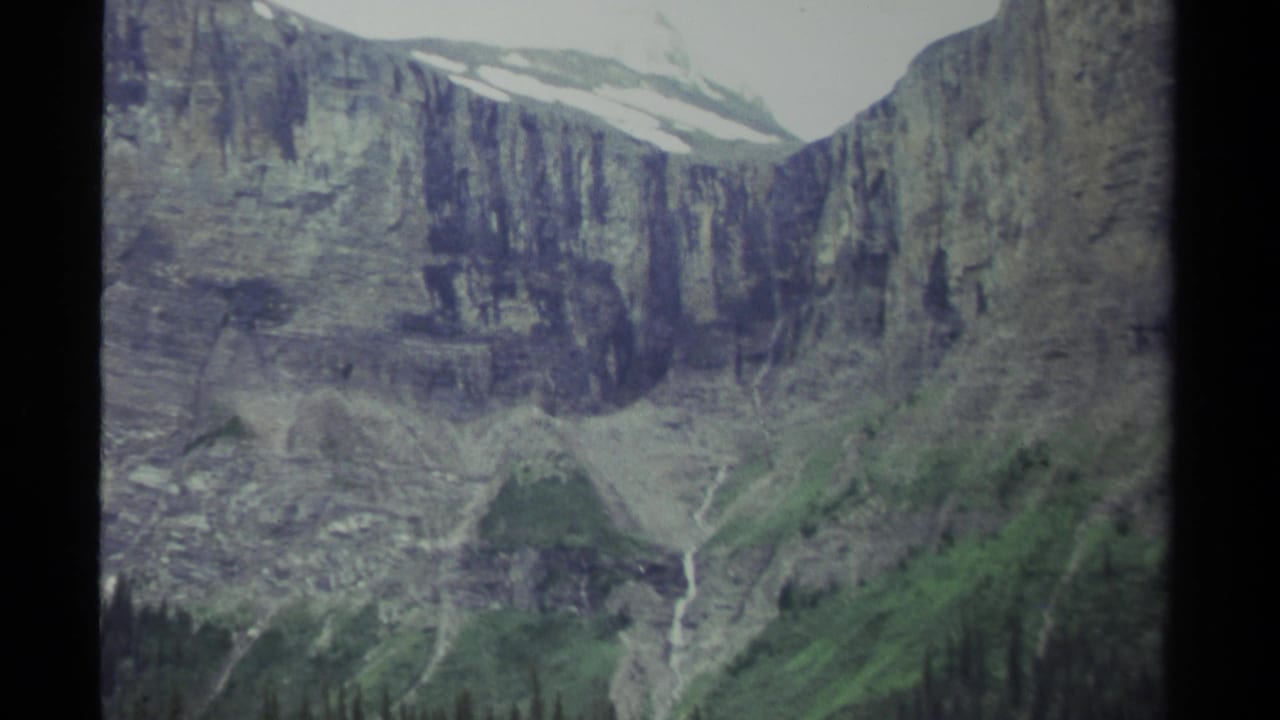 Rocky cliff face with a thin waterfall and green slopes below an overcast sky.