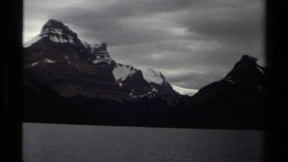 1979 8mm British Columbia landscape frame with a dark lake, snow-dusted rocky peaks, and heavy cloud cover.