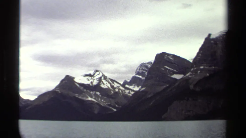 1979 British Columbia 8mm frame showing snow-capped mountain peaks across still water beneath an overcast sky.