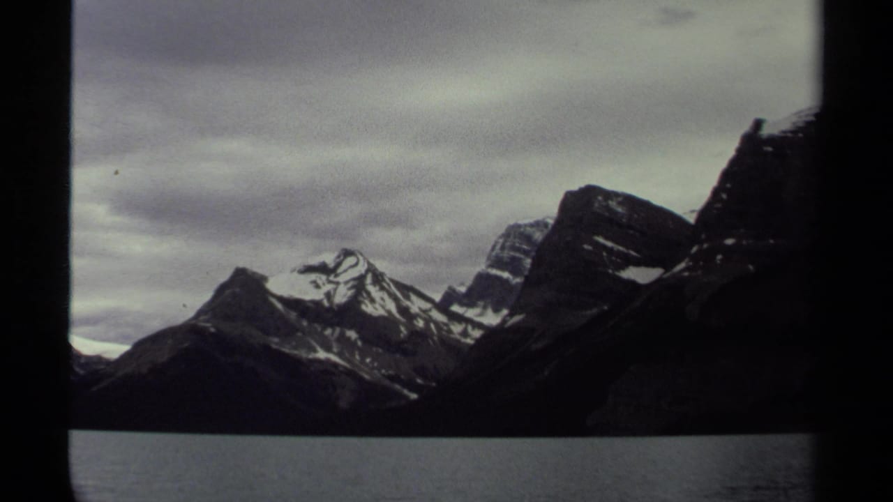 Dark mountain range with snowy peaks seen across water beneath clouds.