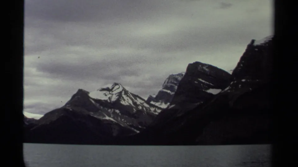British Columbia mountain range across water, with snowy ridges and moody cloud cover in a 1979 home-movie frame.
