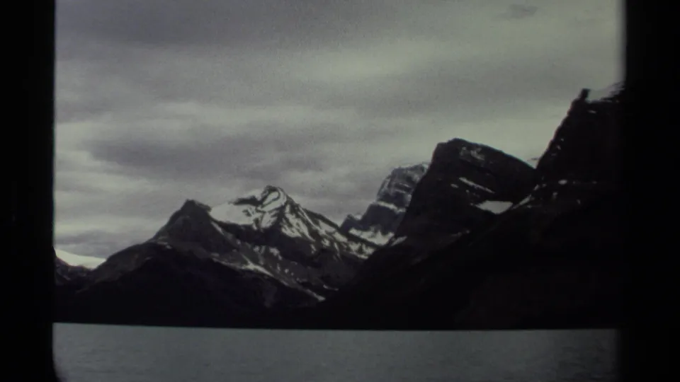 1979 British Columbia 8mm view of snow-streaked peaks, dark foreground slopes, and a calm waterfront under gray clouds.