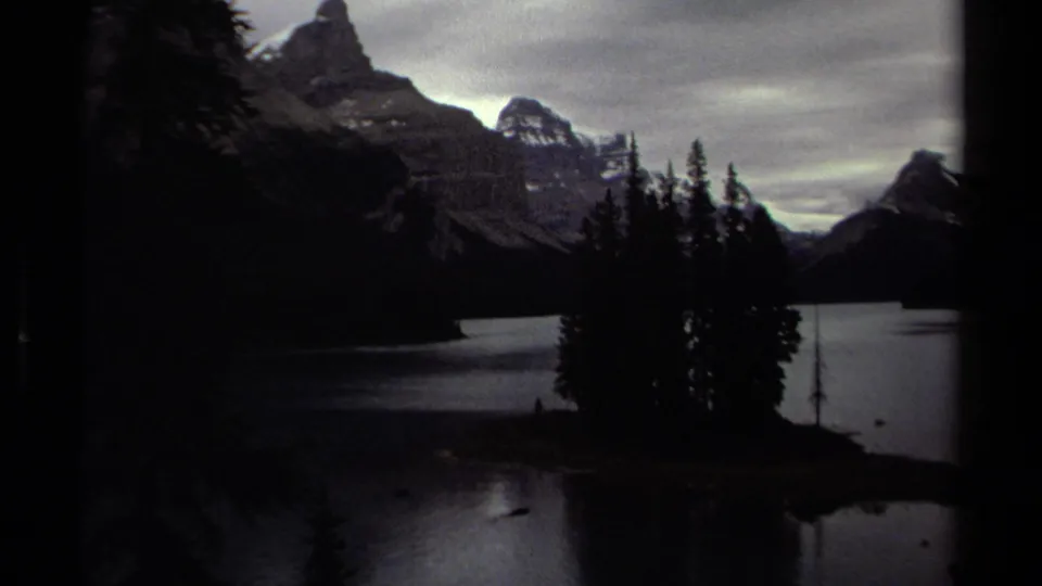 1979 British Columbia lake scene with a small wooded island in front of distant mountain peaks and cloudy sky.