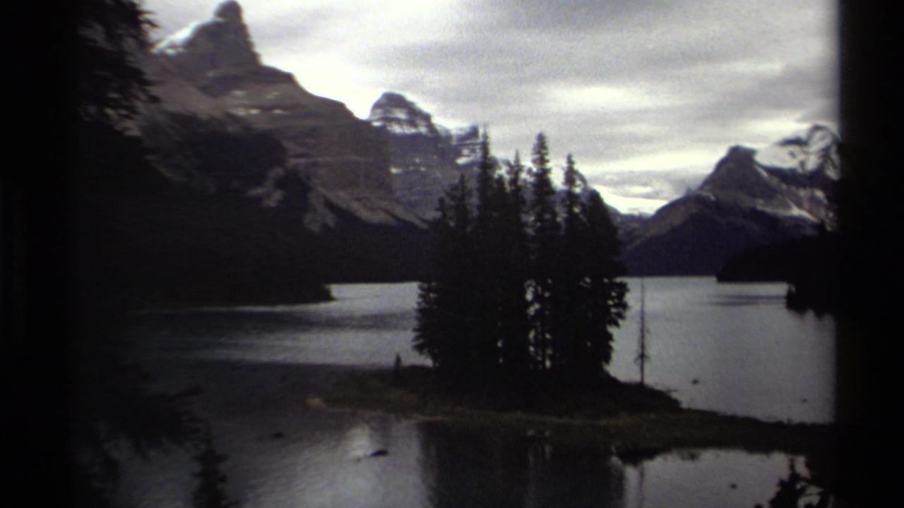Snow-capped mountains rise beyond a calm lake with dark evergreen trees and a small island in the foreground.