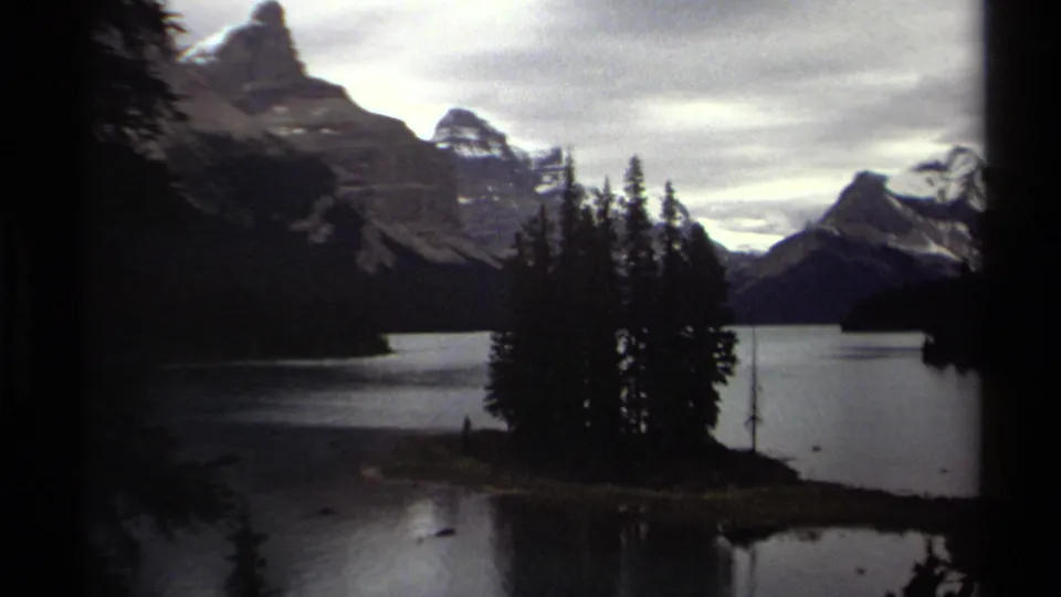1979 8mm home-movie frame from British Columbia showing a calm lake, evergreen island, and snow-capped mountains under an overcast sky.