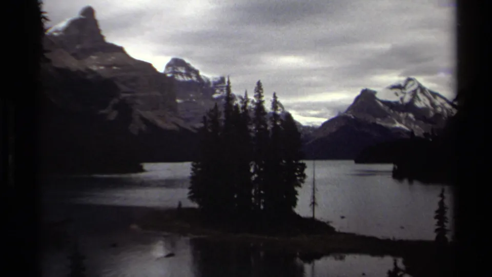 British Columbia 1979 8mm frame showing a wooded island on a calm lake with snow-capped mountains and cloudy sky beyond.