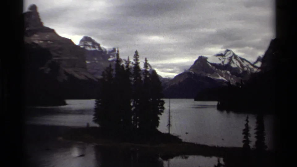 A 1979 British Columbia home-movie frame with evergreen trees, still lake water, dark mountain slopes, and snow-capped peaks in soft focus.
