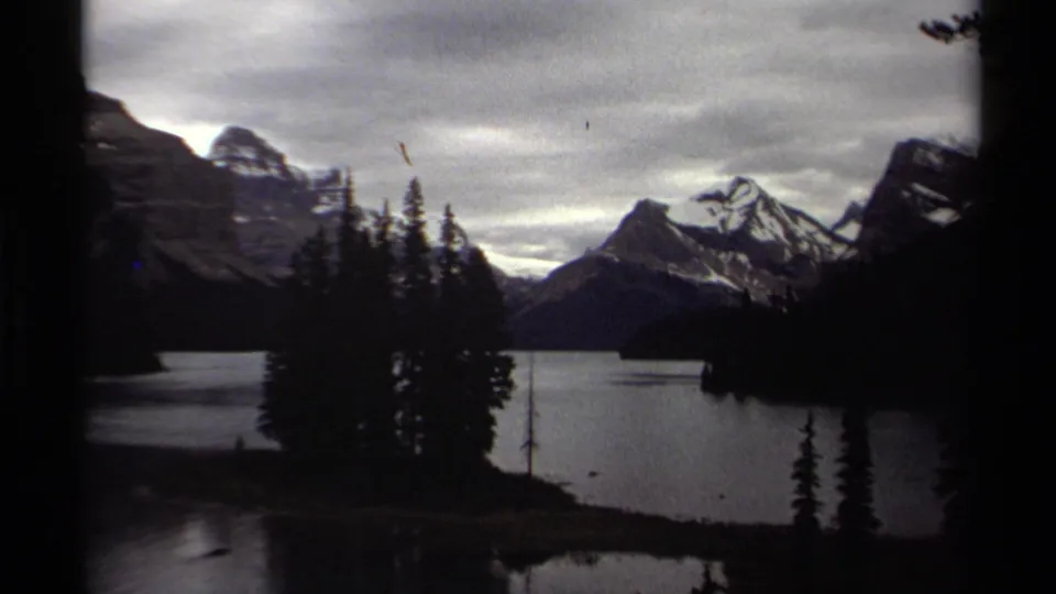 British Columbia 1979 8mm frame with heavy edge shadowing around a calm lake, evergreen island, and snow-capped mountain range.