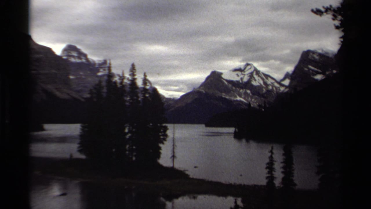 Mountain lake with snow-capped peaks, evergreen silhouettes, cloudy sky, and dark film edges.