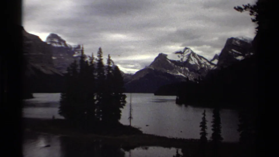 1979 British Columbia 8mm frame showing a mountain lake, snow-capped peaks, evergreens, heavy cloud cover, and dark gate-edge shadows.