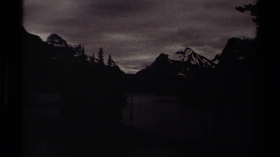 Dark 8mm frame from British Columbia showing a mountain lake and snow-capped peaks partly obscured by tree silhouettes and underexposure.