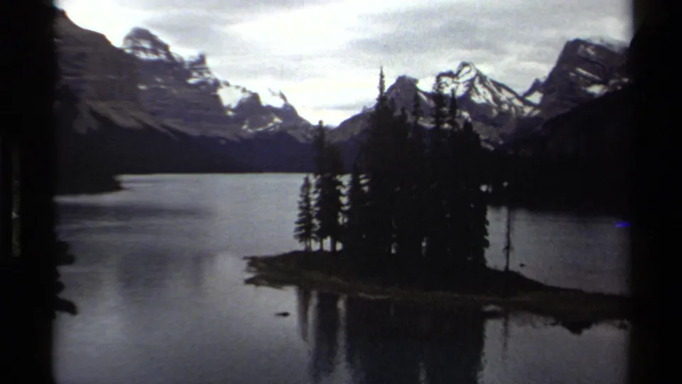 1979 British Columbia 8mm view of a mountain lake with a small evergreen-covered island or shoreline point below snow-capped peaks.