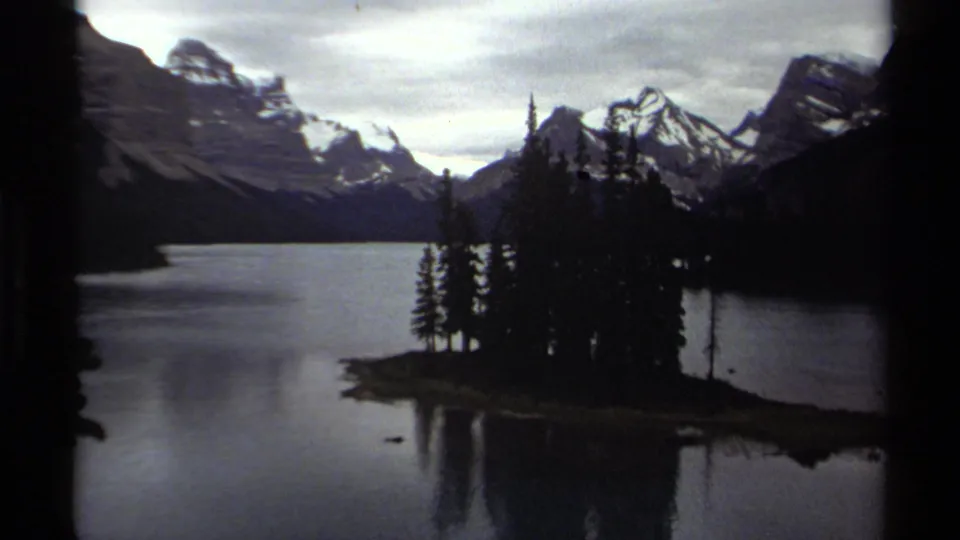British Columbia 8mm frame showing a calm lake, evergreen island or point, distant snow-capped mountains, overcast sky, and visible film-frame border.