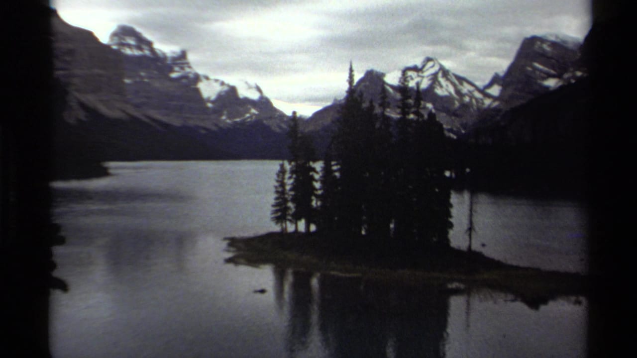 1979 8mm view of a British Columbia mountain lake with a small evergreen island, dark shoreline, and snow-streaked peaks.