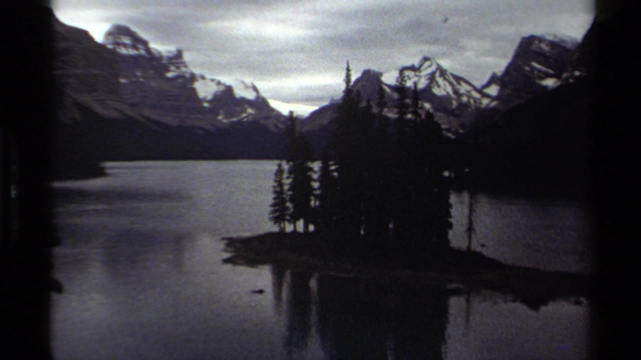Dark mountain lake scene with evergreen island, distant peaks, and cloudy sky.