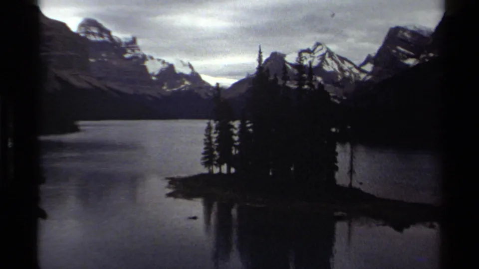 British Columbia home-movie frame showing a lake, tree-covered island, and distant snow-streaked mountains in low light.