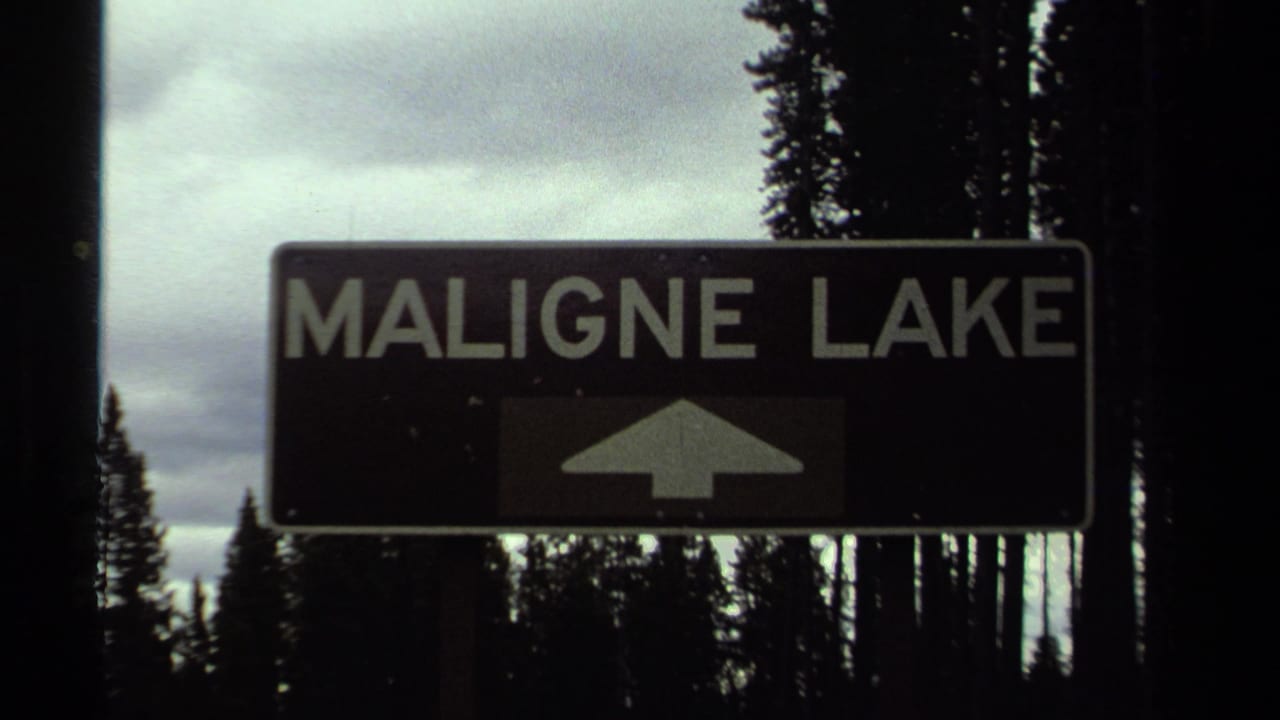 Vintage 8mm frame of the Maligne Lake sign, with the arrow panel partly dark and conifers silhouetted behind it.