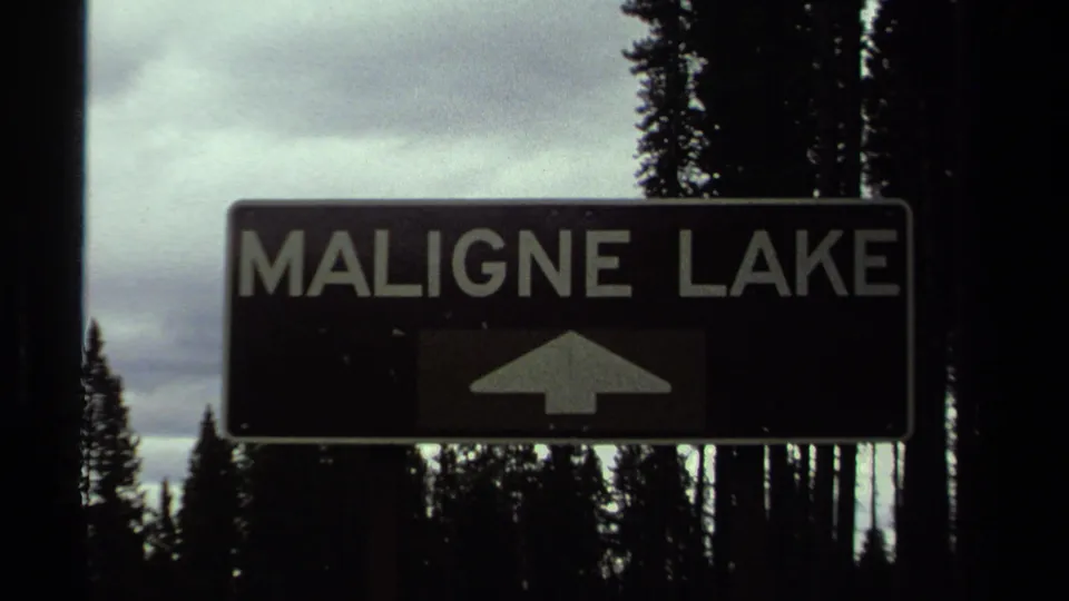 Cataloged 1979 Canadian home-movie frame holding on the Maligne Lake sign, with evergreens and cloudy light behind it.