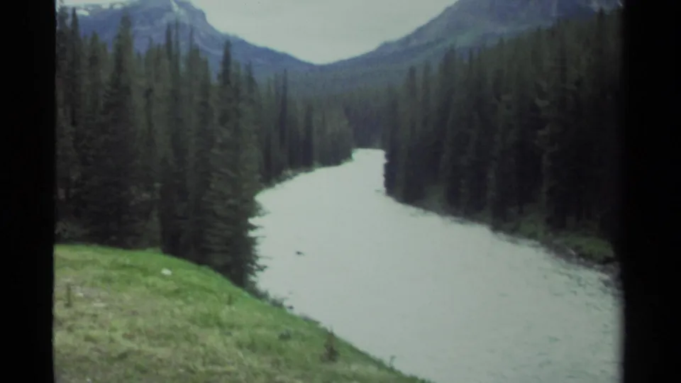 Scenic 8mm travel frame of a pale river or lake channel running through dense forest toward distant mountain slopes.