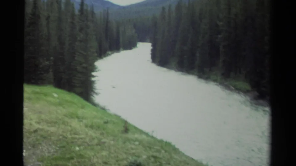 1979 British Columbia 8mm frame of a milky river curving through dense evergreen forest beneath soft overcast light.