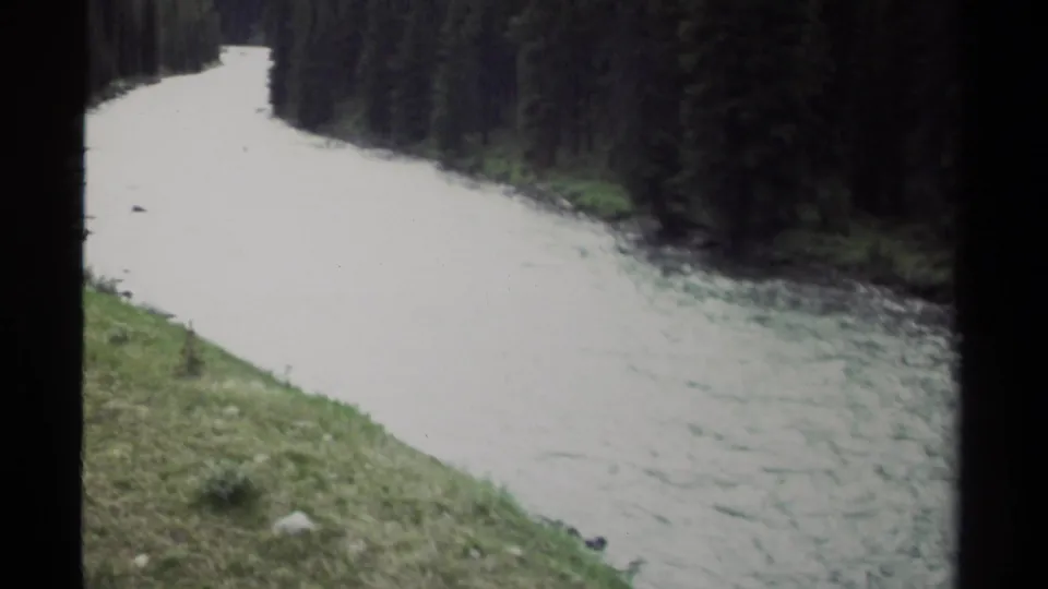 1979 British Columbia 8mm frame showing the river curving beside dark evergreens and a green foreground slope.