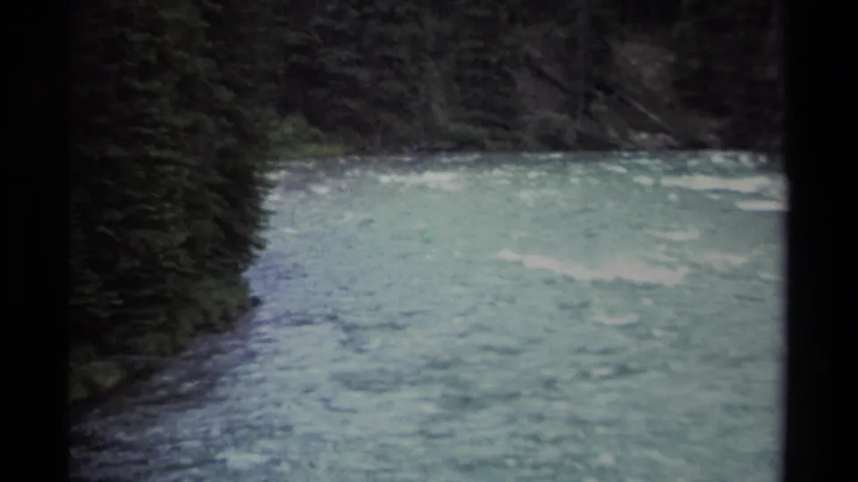 1979 British Columbia 8mm close view of blue-green moving river water below a dark evergreen shoreline.