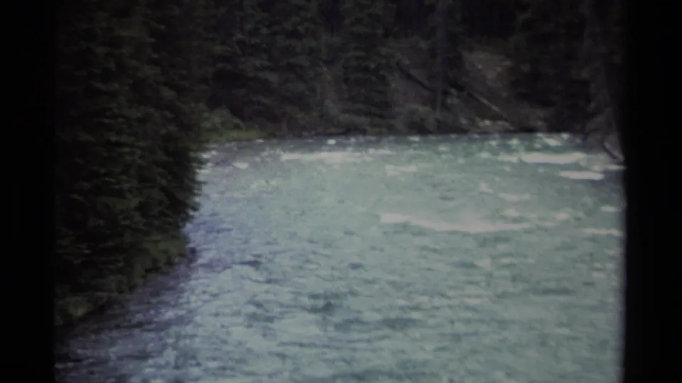 1979 British Columbia 8mm frame showing a pale rushing river curving through dense evergreen forest, bordered by the dark edges of the film scan.